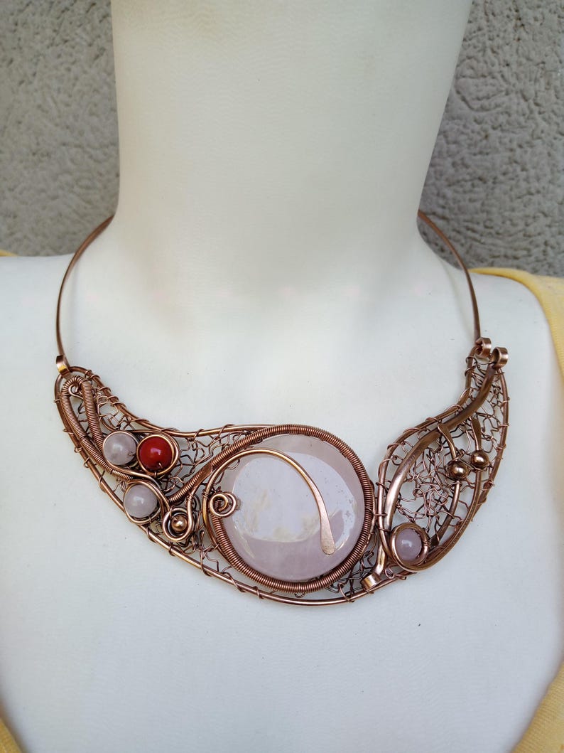 May include: A wire-wrapped copper necklace featuring a large, round, pale pink stone centerpiece. The design includes intricate copper wire filigree, smaller light pink beads, and a single vibrant red bead, all set on a rigid copper collar.