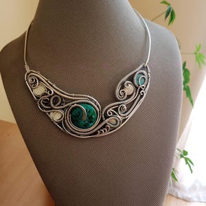 May include: A silver-coloured necklace with detailed wirework. It features a large, round, green stone at the centre, encircled by swirling wire patterns. Smaller white and teal stones are also included. The necklace is displayed on a mannequin, showcasing its intricate design.
