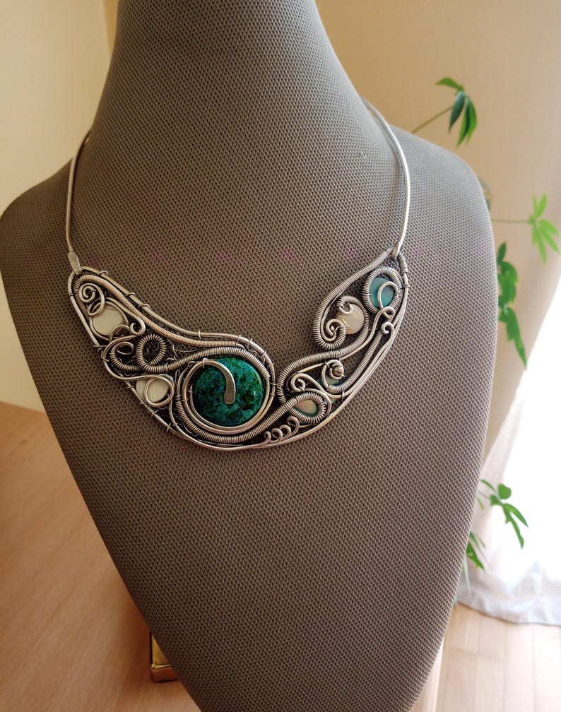 May include: A silver-coloured necklace with detailed wirework. It features a large, round, green stone at the centre, encircled by swirling wire patterns. Smaller white and teal stones are also included. The necklace is displayed on a mannequin, showcasing its intricate design.