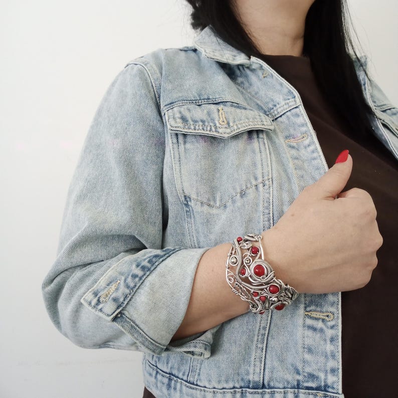 May include: A silver-toned cuff bracelet with red gemstone accents. The bracelet features an intricate wire design with multiple circular and oval red stones. The person is wearing a light blue denim jacket and a brown top.