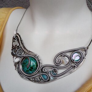 May include: A handcrafted silver wire necklace featuring a large turquoise stone and smaller blue and iridescent stones. The necklace has a unique, flowing design with intricate wirework and is displayed on a mannequin.