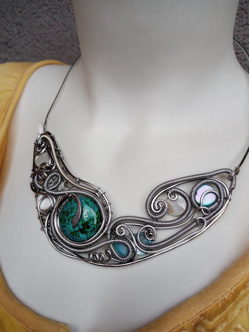 May include: A handcrafted silver wire necklace featuring a large turquoise stone and smaller blue and iridescent stones. The necklace has a unique, flowing design with intricate wirework and is displayed on a mannequin.