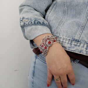May include: Silver-toned cuff bracelet with red gemstone accents. The openwork design features swirling patterns and circular red stones. The bracelet is worn on a wrist, paired with a denim jacket and jeans.