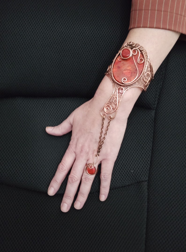 Pu&ograve; includere: Un braccialetto in filo di rame con una grande pietra preziosa rossa e un anello abbinato. Il braccialetto ha una catena delicata che collega il braccialetto all'anello.