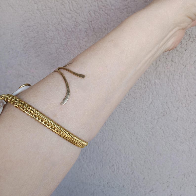 Op de afbeelding: Een goudkleurige armmanchet en bovenarmband. De armmanchet heeft een geweven ontwerp en de bovenarmband heeft een gebogen, open ontwerp. De sieraden worden om de arm van een persoon gedragen.