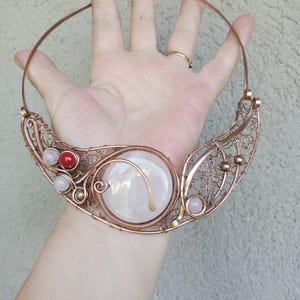 May include: A copper wire collar necklace featuring a large central iridescent cabochon, intricate woven wire mesh detailing, and small accent beads in red and pale pink. The design includes a rigid circular neck wire and decorative metal coils.