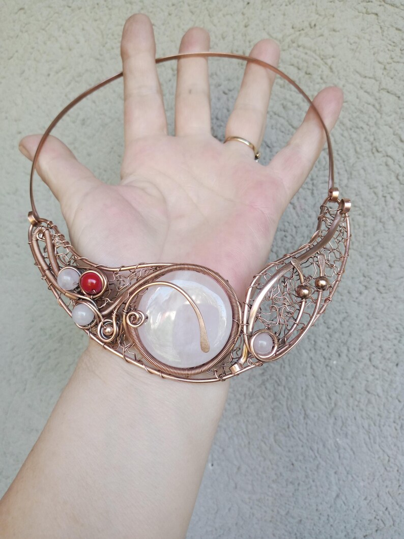 May include: A copper wire collar necklace featuring a large central iridescent cabochon, intricate woven wire mesh detailing, and small accent beads in red and pale pink. The design includes a rigid circular neck wire and decorative metal coils.