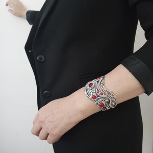 May include: A silver-toned cuff bracelet with intricate wirework and embedded red stones. The bracelet is worn on a wrist, showcasing its ornate design and contrasting colors. The jewelry piece is a statement accessory.