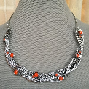 May include: A silver wire necklace with orange beads. The necklace is in the shape of a curved line with swirls and loops. The beads are scattered throughout the necklace.