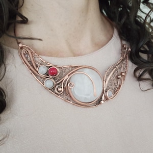 May include: A wire-wrapped copper necklace featuring a large, round, translucent white stone as the centerpiece. The design incorporates intricate copper wire filigree, two smaller translucent white beads, and one vibrant red bead on the left side.