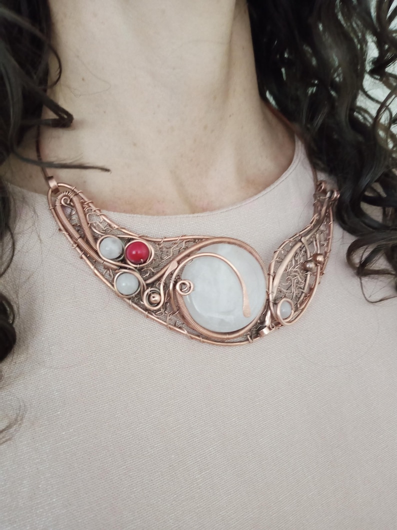 May include: A wire-wrapped copper necklace featuring a large, round, translucent white stone as the centerpiece. The design incorporates intricate copper wire filigree, two smaller translucent white beads, and one vibrant red bead on the left side.