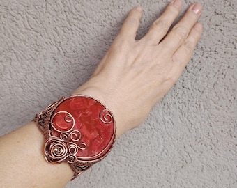 May include: A wide cuff bracelet featuring a large, circular, textured red stone centerpiece. The stone is encased in intricate, swirling copper wirework that forms decorative loops and coils around the edges of the vibrant red gemstone.