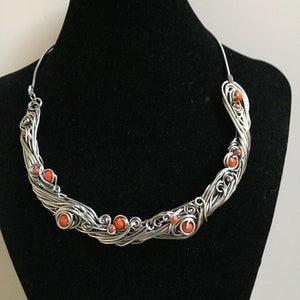 May include: A silver wire necklace with a twisted design and orange beads. The necklace is displayed on a black mannequin bust.
