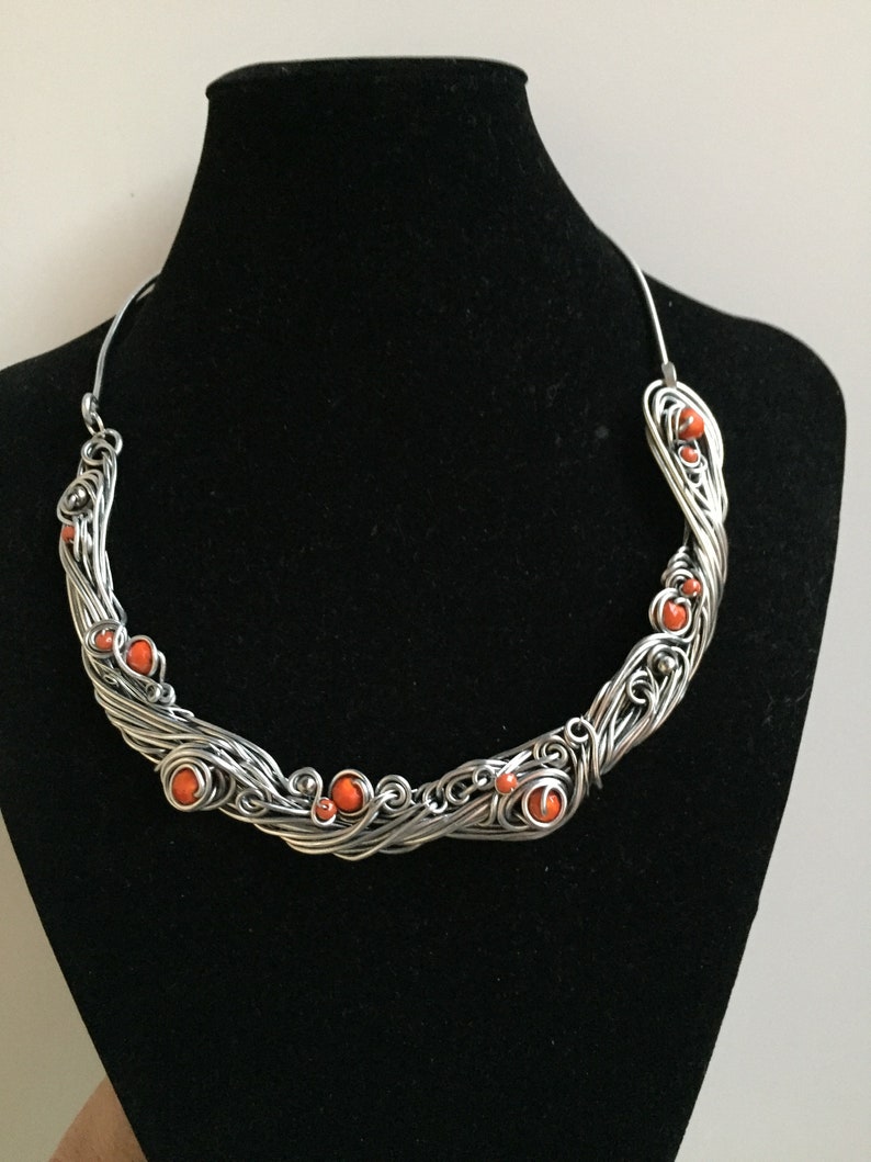 May include: A silver wire necklace with a twisted design and orange beads. The necklace is displayed on a black mannequin bust.