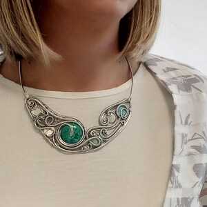 May include: A silver wire-wrapped necklace with a large, round, green stone at its centre. The necklace has a flowing, organic design with smaller turquoise and white accents. The necklace is worn on a person wearing a white shirt and a patterned jacket.