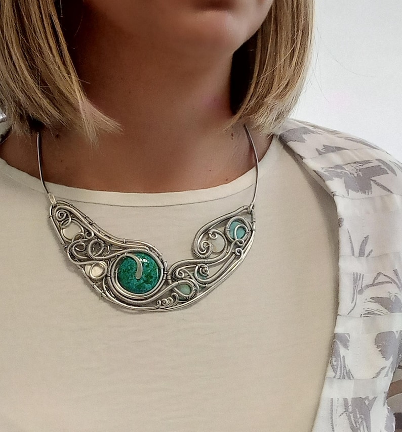 May include: A silver wire-wrapped necklace with a large, round, green stone at its centre. The necklace has a flowing, organic design with smaller turquoise and white accents. The necklace is worn on a person wearing a white shirt and a patterned jacket.