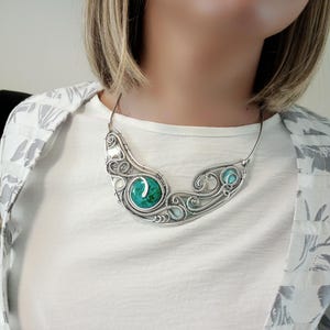 May include: A silver necklace with a curved design, featuring a large, round, turquoise-coloured stone in the centre and smaller turquoise stones on either side. The necklace has an intricate, wire-like pattern and is worn around the neck.