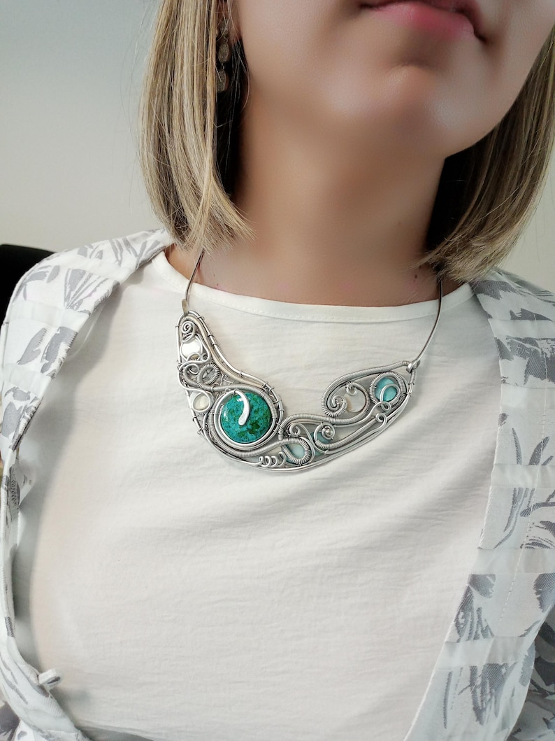 May include: A silver necklace with a curved design, featuring a large, round, turquoise-coloured stone in the centre and smaller turquoise stones on either side. The necklace has an intricate, wire-like pattern and is worn around the neck.