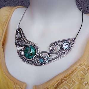 May include: A handcrafted silver wire necklace featuring a large, round, turquoise stone and smaller turquoise and white stones. The necklace has a swirling, openwork design with a black cord.