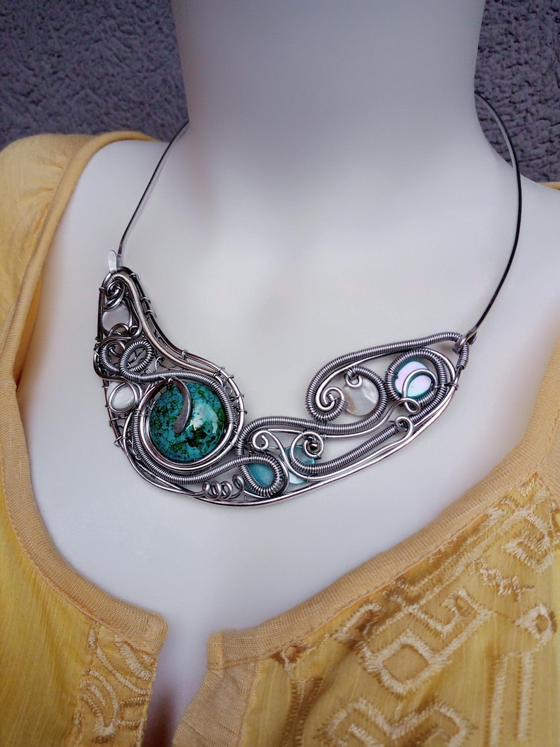 May include: A handcrafted silver wire necklace featuring a large, round, turquoise stone and smaller turquoise and white stones. The necklace has a swirling, openwork design with a black cord.