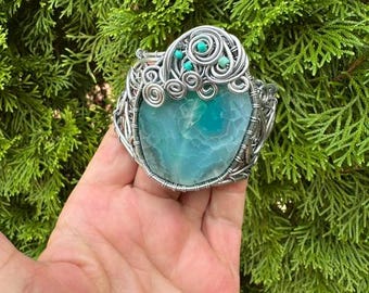 May include: A wire-wrapped cuff bracelet featuring a large, polished, teal-colored agate stone as the centerpiece. The silver-toned metal wire is intricately coiled around the stone and accented with small, round, bright turquoise beads.