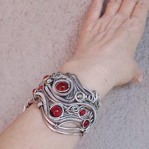 May include: A silver-toned wire-wrapped cuff bracelet with red gemstone accents. The bracelet features a complex, swirling design with multiple red stones of varying sizes. The cuff is worn on a wrist, showcasing the intricate craftsmanship and bold color contrast.