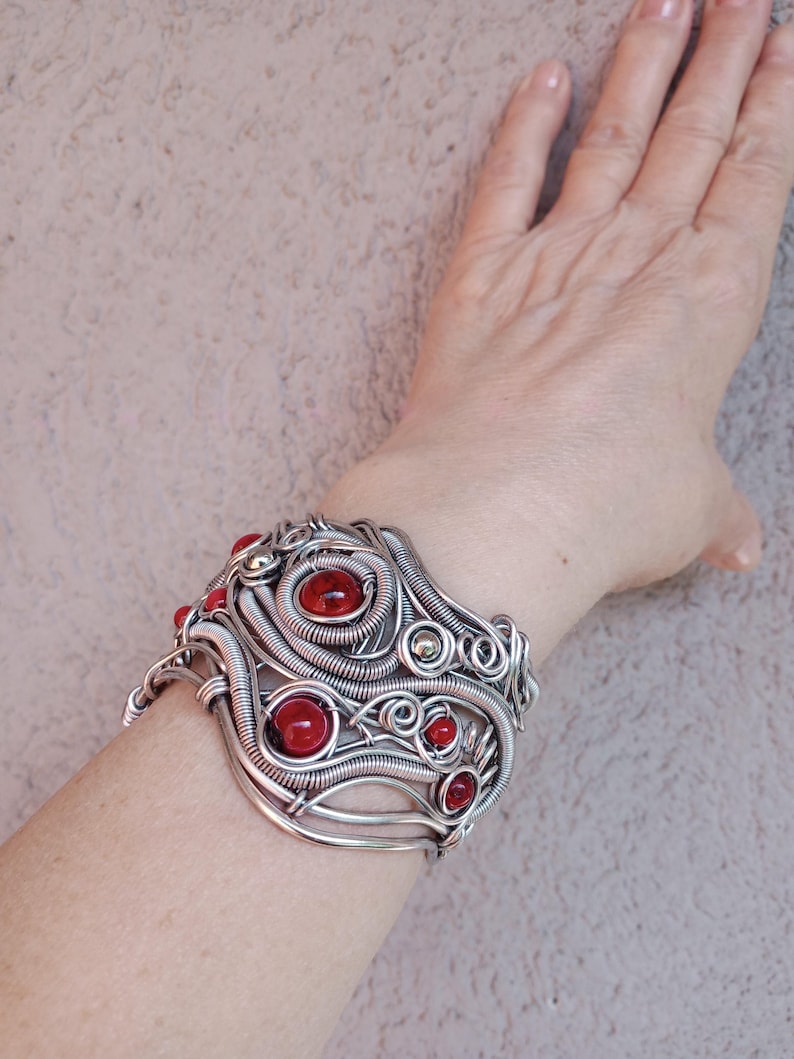 May include: A silver-toned wire-wrapped cuff bracelet with red gemstone accents. The bracelet features a complex, swirling design with multiple red stones of varying sizes. The cuff is worn on a wrist, showcasing the intricate craftsmanship and bold color contrast.