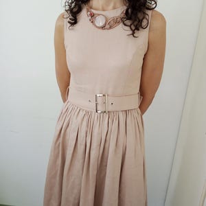 May include: A sleeveless beige dress with a round neckline and a matching wide belt featuring a silver-toned rectangular buckle. The dress has a fitted bodice and a pleated skirt. A wire-wrapped necklace with a central pale stone is worn.