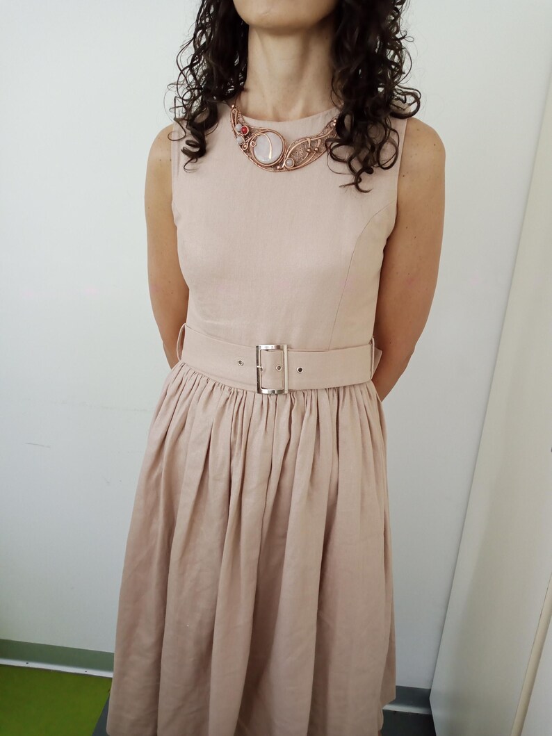 May include: A sleeveless beige dress with a round neckline and a matching wide belt featuring a silver-toned rectangular buckle. The dress has a fitted bodice and a pleated skirt. A wire-wrapped necklace with a central pale stone is worn.