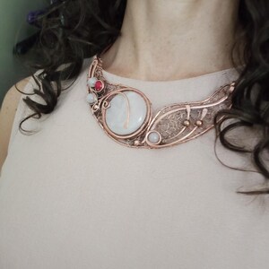May include: A copper wire-wrapped statement necklace featuring a large, round, translucent white stone centerpiece, accented with smaller white and red beads, intricate metal filigree detailing, and a curved, open-work design that rests against the collarbone.