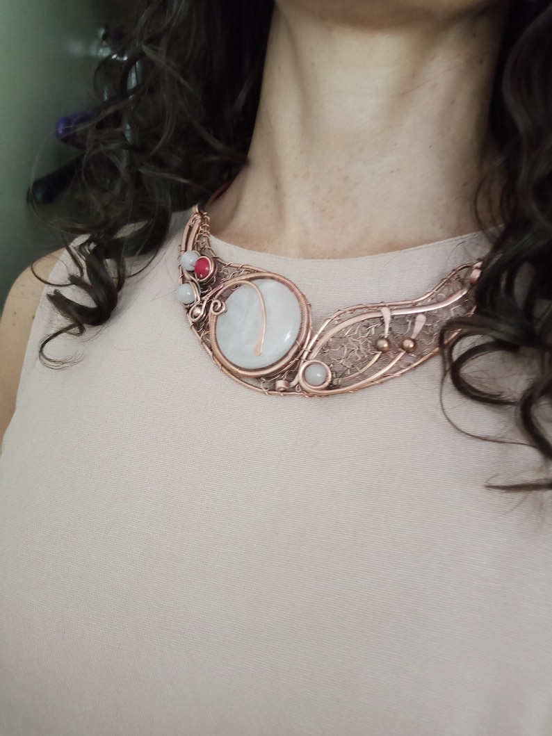 May include: A copper wire-wrapped statement necklace featuring a large, round, translucent white stone centerpiece, accented with smaller white and red beads, intricate metal filigree detailing, and a curved, open-work design that rests against the collarbone.