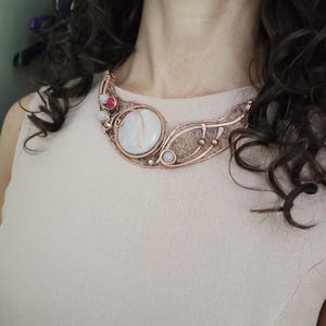 May include: A copper wire-wrapped statement necklace featuring a large, round, milky white central stone. The intricate metalwork includes delicate filigree patterns, small spherical copper accents, and two smaller round stones in white and reddish-pink.