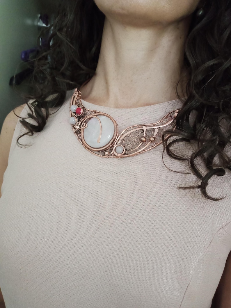 May include: A copper wire-wrapped statement necklace featuring a large, round, milky white central stone. The intricate metalwork includes delicate filigree patterns, small spherical copper accents, and two smaller round stones in white and reddish-pink.