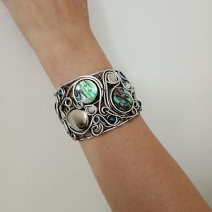 Op de afbeelding: Een brede armband gemaakt van zilverdraad in een wervelend, ingewikkeld patroon. Het ontwerp bevat twee ronde, iriserende inlays van abalone-schelp, een gladde, zilverkleurige metalen kraal en verschillende kleine, donkerblauwe, bolvormige accenten.