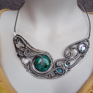 May include: A handcrafted silver wire necklace with a large, round, turquoise stone. The necklace has intricate wirework with smaller stones and iridescent accents. The necklace is displayed on a mannequin.