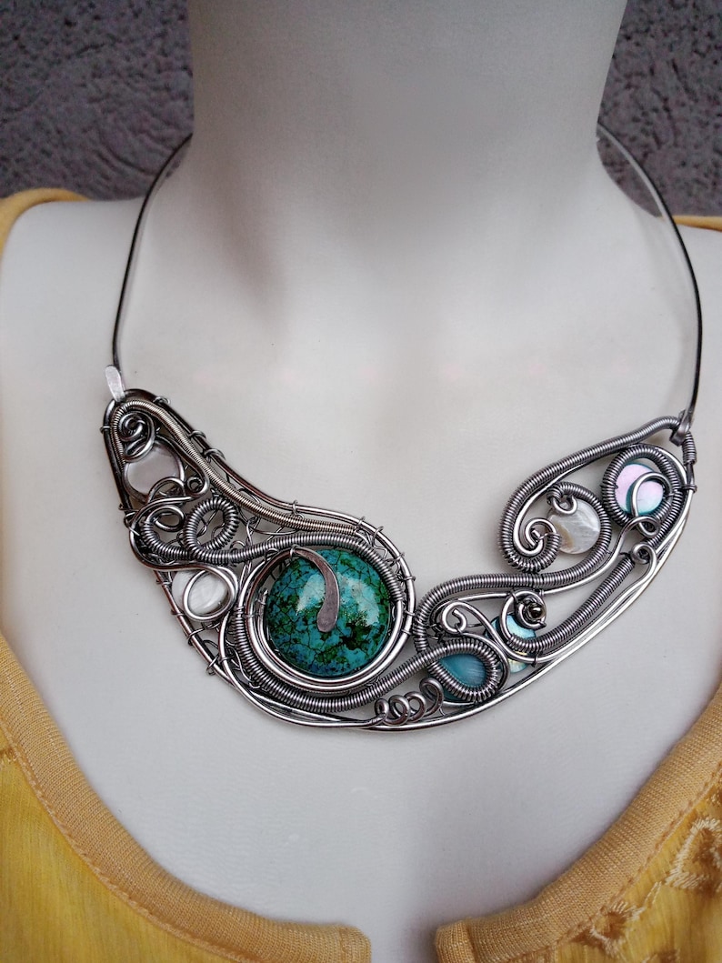 May include: A handcrafted silver wire necklace with a large, round, turquoise stone. The necklace has intricate wirework with smaller stones and iridescent accents. The necklace is displayed on a mannequin.