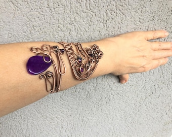 May include: A wide cuff bracelet crafted from copper wire, featuring intricate swirling patterns and woven metal details. The design incorporates a large, smooth, deep purple stone and several smaller, round, dark purple beads integrated into the wirework.