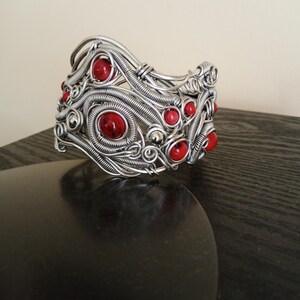 May include: A silver-toned wire-wrapped bracelet with red gemstone accents. The bracelet features intricate wirework with spiraling and woven patterns, and is adorned with multiple round, red gemstones.