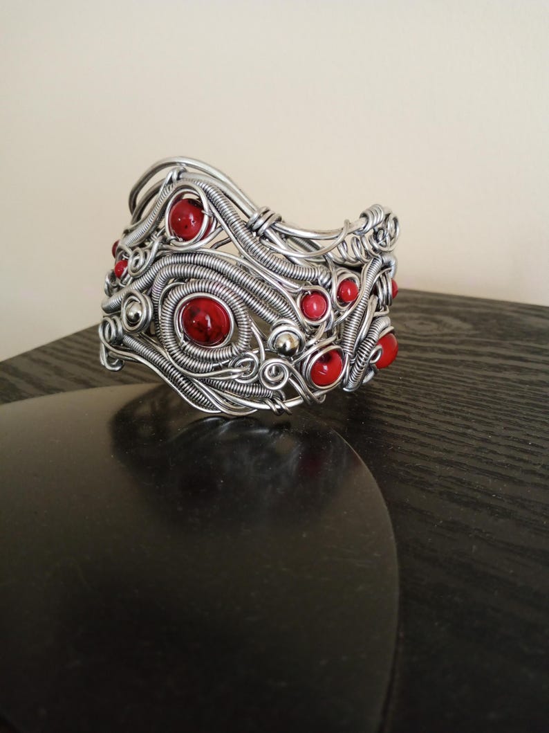 May include: A silver-toned wire-wrapped bracelet with red gemstone accents. The bracelet features intricate wirework with spiraling and woven patterns, and is adorned with multiple round, red gemstones.