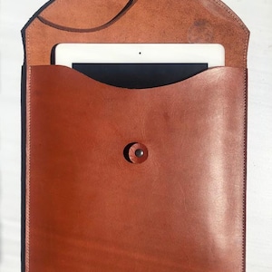 Puede incluir: Una funda para tablet de cuero marrón con cierre de solapa y botón. La funda tiene una correa que se puede usar para llevarla.