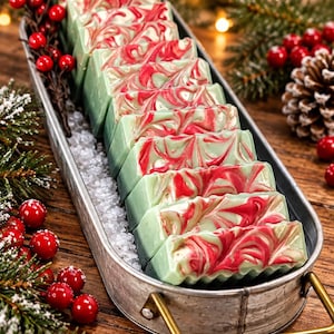 May include: A row of rectangular soap bars with a green and red marbled swirl pattern, arranged inside a galvanized metal tray with gold-toned handles, resting on a bed of white salt crystals.