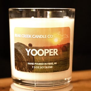Può includere: Una candela in vetro trasparente con cera bianca e coperchio in legno. L'etichetta mostra un campo al tramonto con balle di fieno, il testo BEAR CREEK CANDLE CO, YOOPER, HAND POURED IN FIBRE, MI, 7.5 oz soy blend e la mappa del Michigan.
