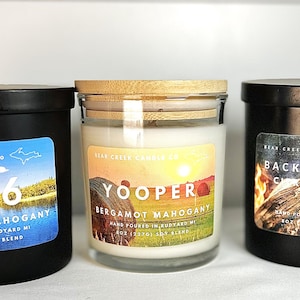 May include: Three black glass candle jars with wooden lids. The labels on the jars read "906 Cedar Oak Mahogany", "Yooper Bergamot Mahogany", and "Back at Camp Citrus & Spice".