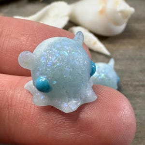 May include: A close-up of two small, light blue, glittery resin sculptures. Each has a rounded body, two small ears, and a turquoise dot on each side. The sculptures are on a finger, with a blurred white seashell in the background.