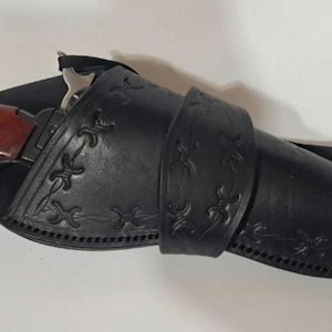 May include: A black leather holster featuring decorative embossed patterns and stitched edges, containing a firearm with a polished reddish-brown wooden grip. The holster is secured with a matching leather strap across the center section.