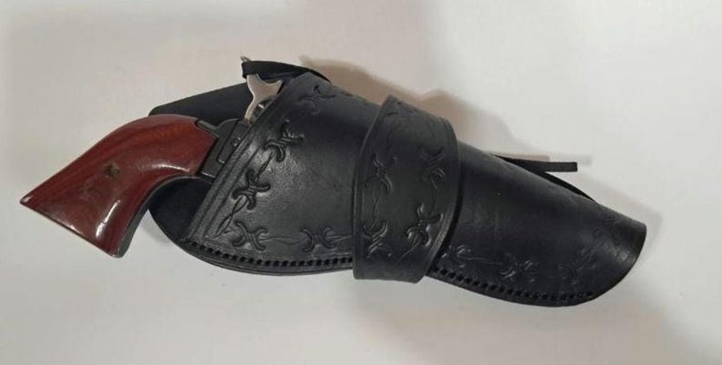 May include: A black leather holster featuring decorative embossed patterns and stitched edges, containing a firearm with a polished reddish-brown wooden grip. The holster is secured with a matching leather strap across the center section.