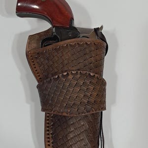 May include: A brown leather holster with a woven design, holding a revolver with a dark wooden grip. The holster has a belt loop and a strap. The revolver is partially visible.