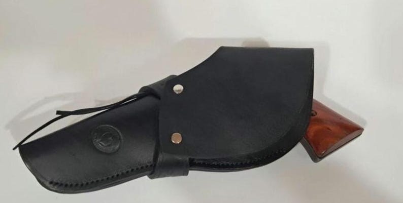 May include: A black leather holster featuring a curved flap secured with two silver-toned metal rivets. The holster has a circular embossed logo on the side and a brown wooden handle of a firearm is visible protruding from the open end of the casing.