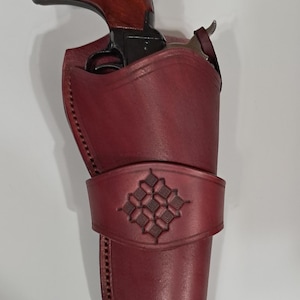 May include: A burgundy leather holster with a decorative embossed design, holding a revolver with a dark wooden handle. The holster features stitched edges and a strap. The revolver is partially visible.