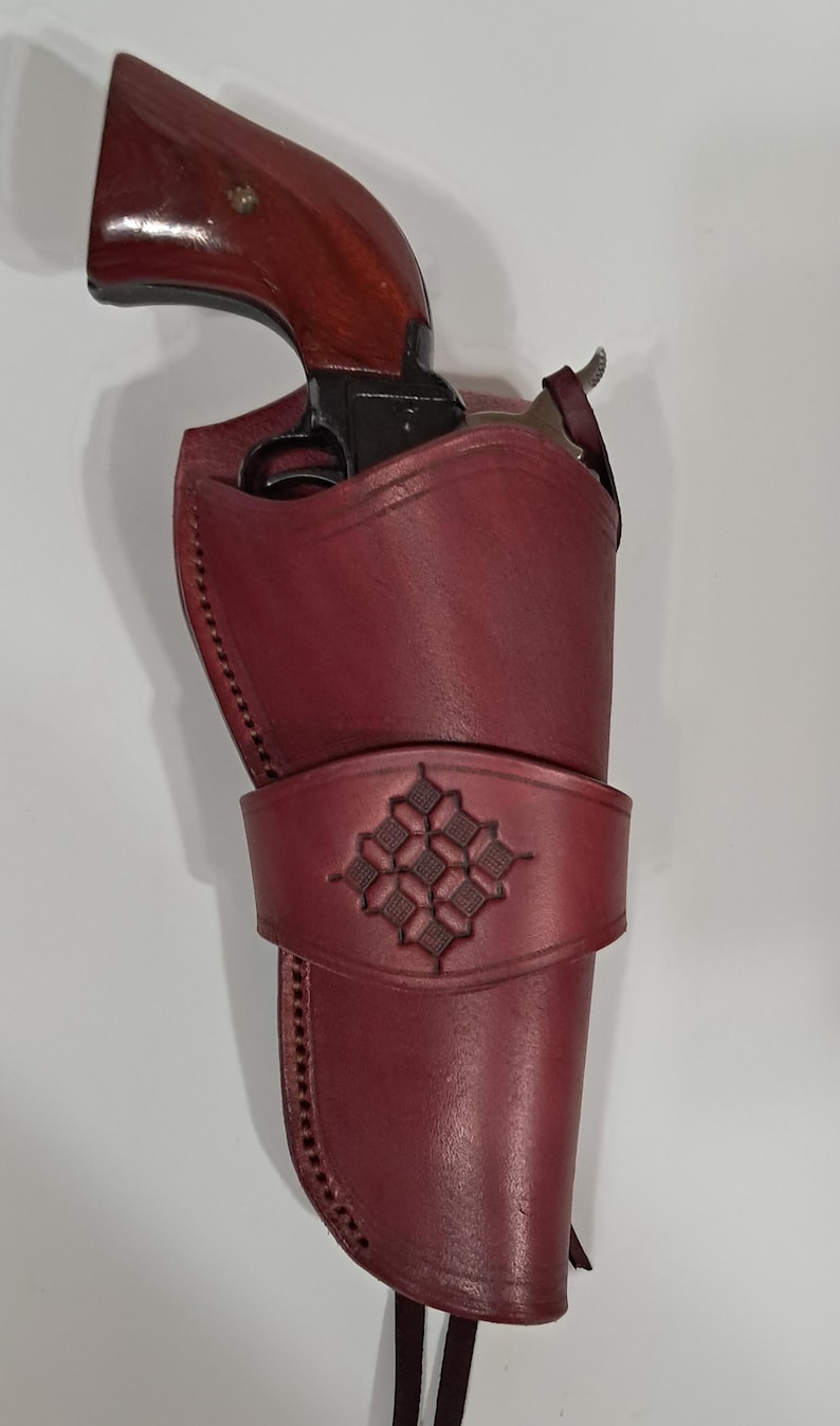 May include: A burgundy leather holster with a decorative embossed design, holding a revolver with a dark wooden handle. The holster features stitched edges and a strap. The revolver is partially visible.